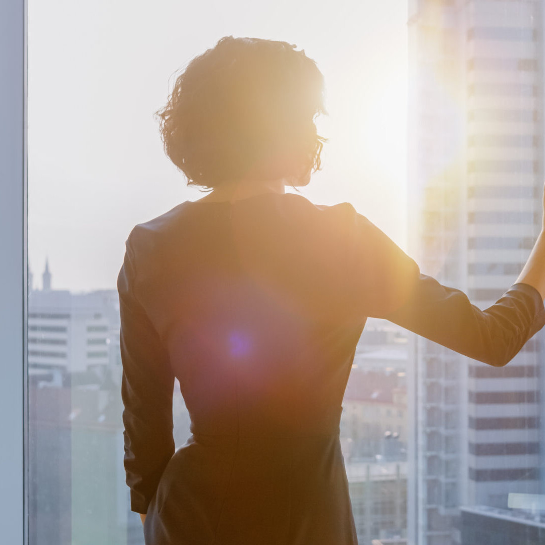 Shot of the Successful Businesswoman in a Striking Black Dress in Her Office Looking out of the Window at Sunset. Modern Business Office with Personal Computer and Big City View.